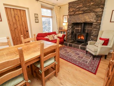 A dining area with a wooden table and chairs next to a sitting area with a red sofa and a wingback chair by a stone fireplace at Nutkin Cottage in Bowness-On-Windermere
