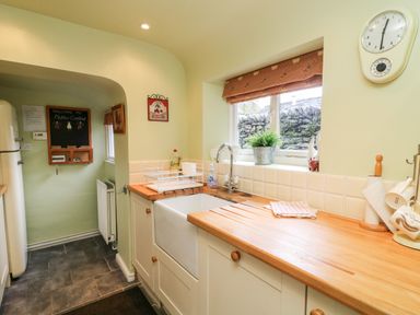 A kitchen with a wooden countertop a white farmhouse sink a window with a patterned blind and a clock on the wall at Nutkin Cottage in Bowness-On-Windermere