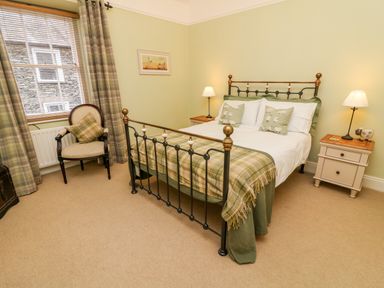 A bedroom with a bed with green plaid blanket two bedside tables with lamps and a chair next to a window at Nutkin Cottage in Bowness-On-Windermere