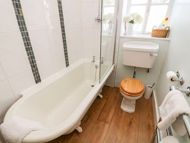 A bathroom with a bathtub a toilet with a wooden seat and a window with a basket and flowers on the sill at Nutkin Cottage in Bowness-On-Windermere