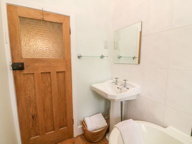 A bathroom with a wooden door a pedestal sink a mirror a wicker basket with towels and a bathtub with a towel at Nutkin Cottage in Bowness-On-Windermere
