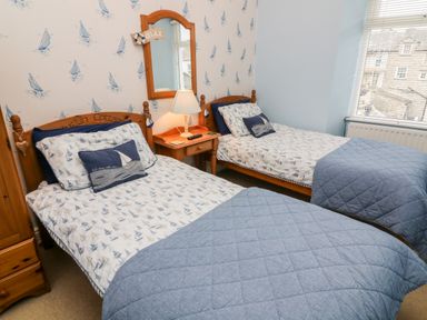 A bedroom with two single beds with boat-themed bedding a wooden nightstand with a lamp and a mirror above at Nutkin Cottage in Bowness-On-Windermere