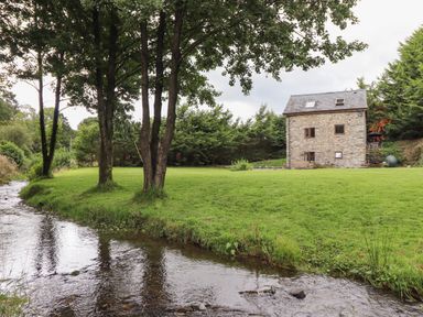 A stone house with a grassy yard and trees next to a stream at Yr Hen Felin in Llanfyllin