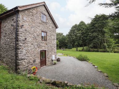 A stone building with small windows and a door surrounded by gravel and grass with trees in the background at Yr Hen Felin in Llanfyllin