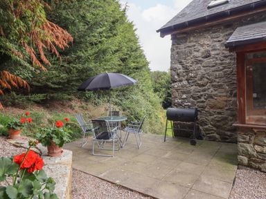 An outdoor patio with a round table and four chairs under a black umbrella next to a stone building and potted red flowers at Yr Hen Felin in Llanfyllin