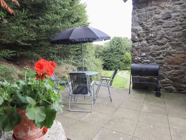 An outdoor patio with a round table under a black umbrella surrounded by four chairs a flower pot with red flowers and a barbecue grill near a stone wall at Yr Hen Felin in Llanfyllin