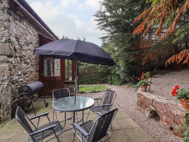 An outdoor patio with a round glass table four chairs and a black umbrella next to a stone wall and a barbecue at Yr Hen Felin in Llanfyllin