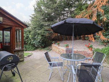 An outdoor patio with a glass table shaded by a black umbrella and surrounded by four chairs next to a barbecue grill and garden area at Yr Hen Felin in Llanfyllin