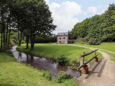 A small stone house surrounded by trees with a stream and a wooden bridge in the foreground at Yr Hen Felin in Llanfyllin
