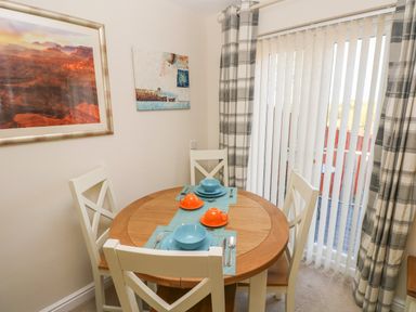 A dining area with a wooden round table set with blue and orange dishes and four white chairs near window with vertical blinds at Ffau'r Ddraig in Cross Hands
