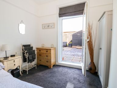 A bedroom with a bedside table lamp an iron towel rack a wooden chest of drawers a wardrobe and a door opening to a patio at Northgate Ground Floor Flat in Hunstanton