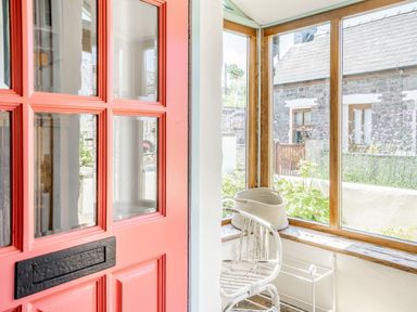 A porch area with a red door white chair and woven basket by windows at Sea Pickle Cottage in Llangwm Pembrokeshire