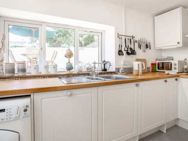 A kitchen with white cabinets wooden countertops a double sink utensils hanging on the wall and a window showing an outdoor patio at Sea Pickle Cottage in Llangwm Pembrokeshire