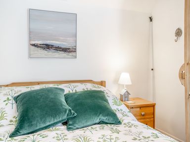 A bedroom with a wooden bed featuring green pillows and floral bedding a nightstand with a lamp and a decorative house and a painting on the wall at Sea Pickle Cottage in Llangwm Pembrokeshire