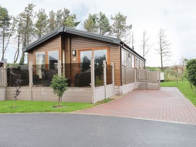 A wooden cabin with glass doors and a fenced porch with chairs beside a paved driveway at Huckleberry in Runswick Bay near Staithes