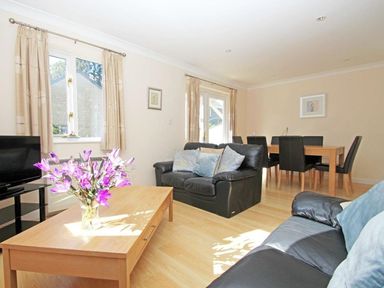 A living room with black leather sofas a wooden coffee table with purple flowers a dining table with black chairs and windows with beige curtains at Waters Edge in Falmouth