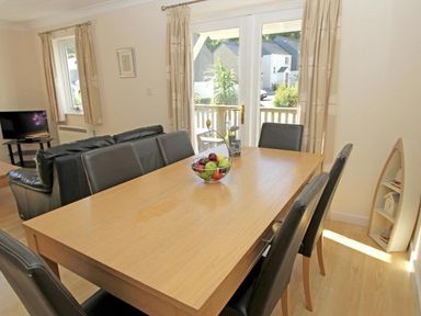 A dining area with a wooden table and six black chairs a fruit basket on the table a black leather sofa a television on a stand and glass doors leading outside at Waters Edge in Falmouth