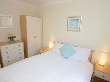 A bedroom with a bed white bedding two bedside tables with lamps a chest of drawers with flowers and a wardrobe at Waters Edge Falmouth