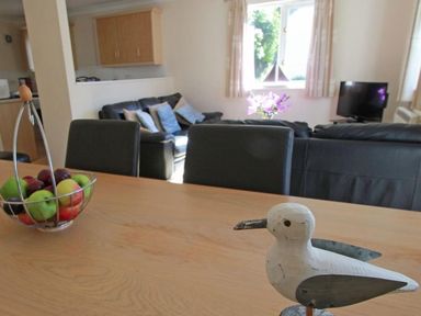 A dining table with a bowl of fruit and a wooden bird decoration with chairs and a living area with sofas and a television at Waters Edge in Falmouth