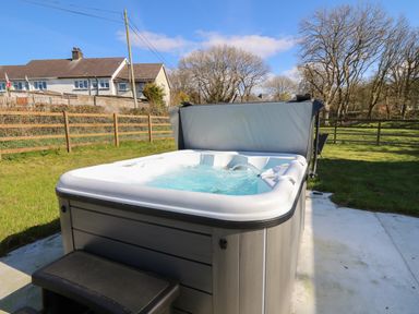 An outdoor hot tub with bubbling water on a concrete patio in a fenced grassy yard at Glyn Cottage in Llanon