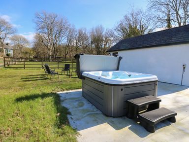 An outdoor area with a hot tub and plastic steps on a concrete patio with chairs on grass behind at Glyn Cottage in Llanon