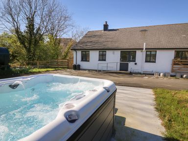 An outdoor hot tub with water jets in front of a white house with a sloped roof and fenced garden at Glyn Cottage in Llanon