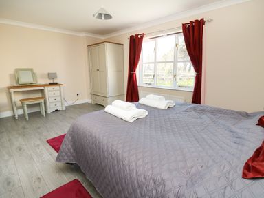 A bedroom with a bed covered with a grey quilt and two white towels on top two red rugs on the floor a cream wardrobe near a window with red curtains and a cream dressing table with a stool and lamp at Glyn Cottage in Llanon