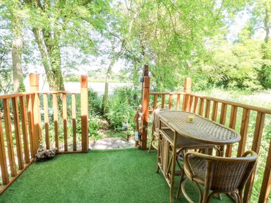 A wooden deck with a wicker table and two wicker chairs surrounded by green trees at Hugatree in Little Dunham near Litcham
