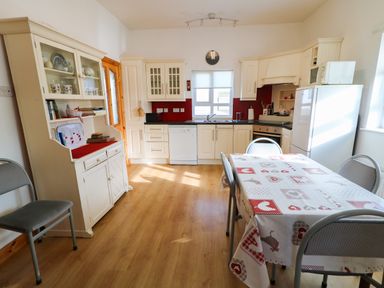 A kitchen with white cabinets and appliances including a dishwasher and refrigerator a table with four chairs and patterned tablecloth a hutch with dishes and books at Ard an Phiobaire in An Luinnigh near Derrybeg County Donegal