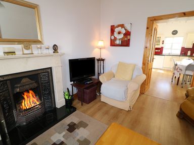 A living room with a fireplace, armchair, television, lamp, and a view into a kitchen at Ard an Phiobaire in An Luinnigh near Derrybeg County Donegal