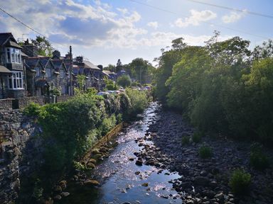 A small river flowing between stone houses with parked cars and trees lining the riverbank at Y Bwthyn in Llanbedr