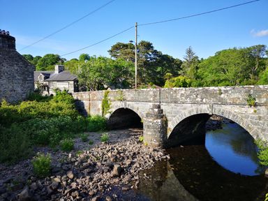 A stone arch bridge over a river with trees and a stone building in the background at Y Bwthyn in Llanbedr