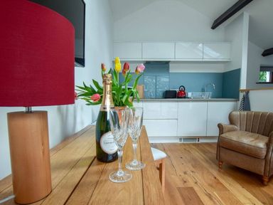 A kitchen area with white cabinets a wooden table with a bottle of champagne and glasses a lamp and tulips and a leather armchair at Old Sawrey Stores in Hawskhead