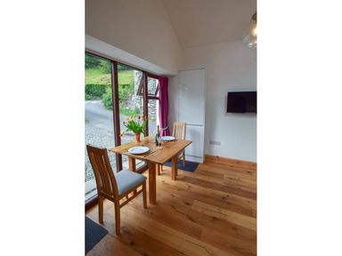 A dining area with a wooden table set for two with a vase of flowers next to large windows at Old Sawrey Stores in Hawskhead