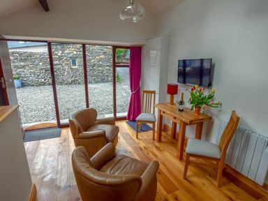 A sitting area with two brown leather chairs two wooden chairs a wooden table with flowers and a lamp and a wall-mounted TV at Old Sawrey Stores in Hawskhead