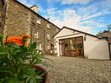A stone building and a white building with glass doors overlooking a gravel courtyard with orange flowers in a pot at Old Sawrey Stores in Hawskhead