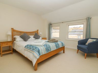 A bedroom with a wooden bed with pillows and towels on it two wooden bedside tables with lamps two windows with curtains and a blue armchair at Kernyk in Perranuthnoe