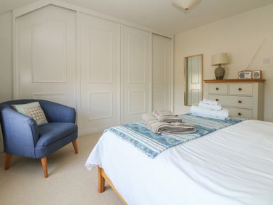 A bedroom with a bed, towels on the bed, a blue armchair, a dresser with a lamp and radio, and a tall mirror at Kernyk in Perranuthnoe