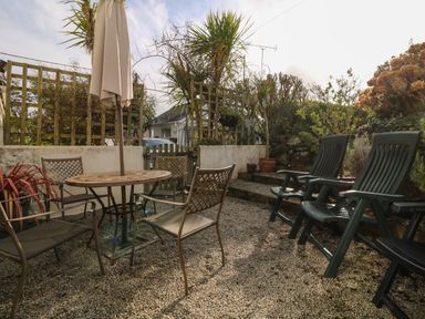 An outdoor patio area with a round table and chairs and plastic armchairs surrounded by plants and greenery at Kernyk in Perranuthnoe