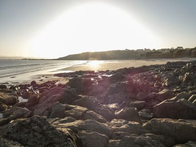 A rocky coastline with a sandy beach and houses on a hill with the sun setting in the background at Kernyk in Perranuthnoe