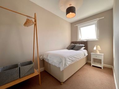 A small bedroom with a bed pillows bedside table with lamp window and a wooden clothes rack with hangers and storage baskets at Kernyk in Perranuthnoe