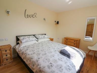 A bedroom with a bed and floral bedding two wooden bedside tables a wooden chest of drawers a mirror on the wall and a white chair at Ploony Hill Cabin in Bleddfa near Knighton