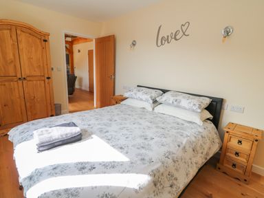 A bedroom with a double bed covered in floral bedding two wooden nightstands a wooden wardrobe and an open door at Ploony Hill Cabin in Bleddfa near Knighton