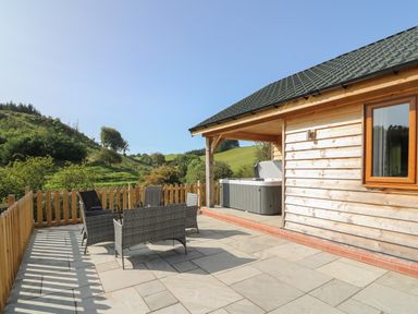 A wooden cabin patio with outdoor chairs and a hot tub overlooking green hills at Ploony Hill Cabin in Bleddfa near Knighton