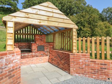 An outdoor brick barbecue area with a wooden slatted roof and surrounding fence at Ploony Hill Cabin in Bleddfa near Knighton