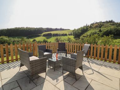 An outdoor patio with five chairs and a glass table with a bottle and two glasses with a wooden fence and green hills in the background at Ploony Hill Cabin in Bleddfa near Knighton