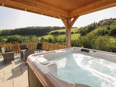 An outdoor patio with a hot tub and seating area overlooking green hills and trees at Ploony Hill Cabin in Bleddfa near Knighton
