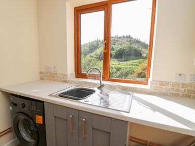 A kitchen sink with grey cabinets and a washing machine under a window overlooking greenery at Ploony Hill Cabin in Bleddfa near Knighton