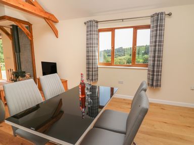 A dining area with a black glass table and gray chairs next to a window with checkered curtains and a television near a wood-burning stove at Ploony Hill Cabin in Bleddfa near Knighton