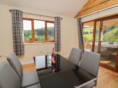 A dining area with a black glass table and four gray chairs next to windows with plaid curtains and a sliding door showing a hot tub outside at Ploony Hill Cabin in Bleddfa near Knighton